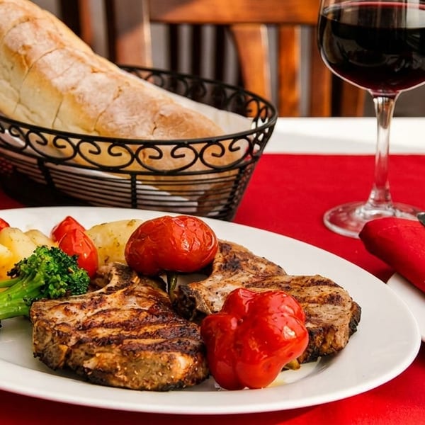 Grilled pork chops with roasted vegetables, bread, and red wine on a red placemat.