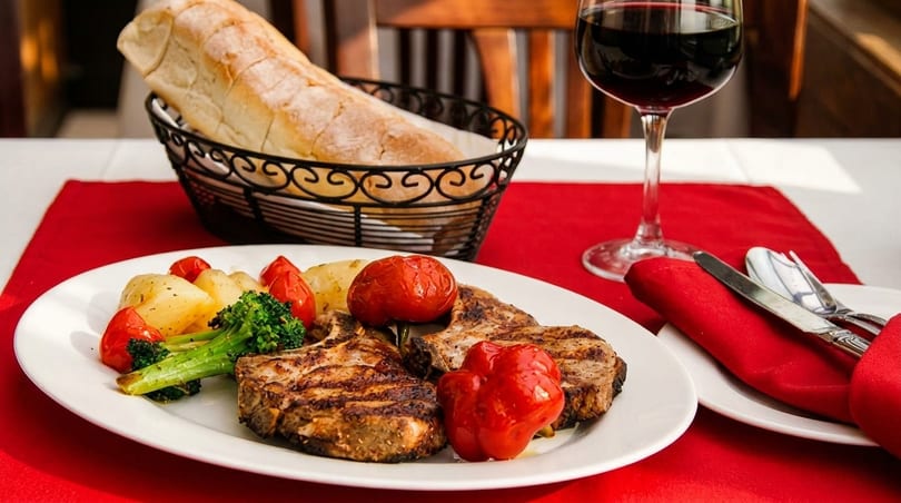 Grilled pork chops with roasted vegetables, bread, and red wine on a red placemat.