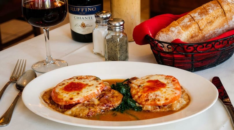Chicken parmesan with melted cheese and tomato, spinach, red wine, and bread on a white tablecloth.