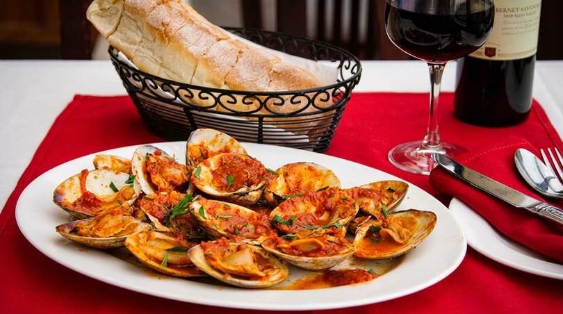 Steamed clams in red sauce on a white plate, with bread, wine, and silverware on a red tablecloth.