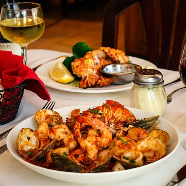 Two plates of seafood, bread, and wine glasses on a white tablecloth in a restaurant setting.