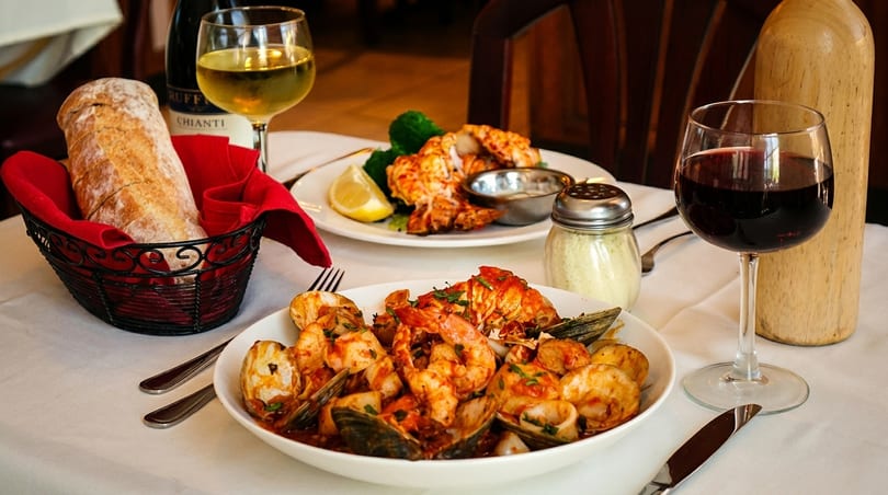 Two plates of seafood, bread, and wine glasses on a white tablecloth in a restaurant setting.